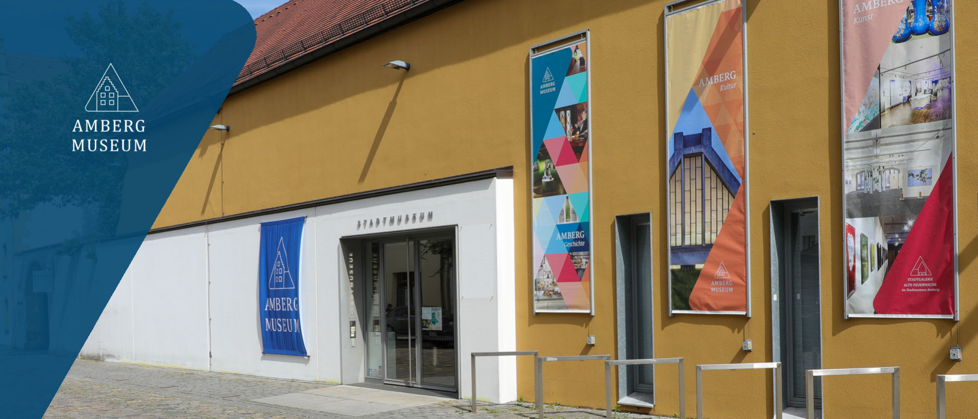 Außenansicht vom Stadtmuseum Amberg mit Eingangstür, einer bunten Fahne links und drei bunten Bannern rechts davon.
