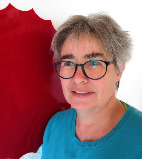 Brustbild der Künstlerin Gisela Hoffmann vor einer weißen Wand mit einem roten Kreis. Sie hat kurzes graues Haar, trägt eine Brille mit schwarzem Gestell und ein türkis-blaues T-shirt.
