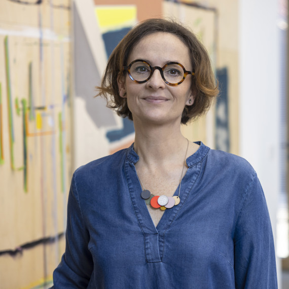 Brustbild der Künstlerin Sandra Tröger vor einem bunten Kunstwerk. Sie trägt das braune Haar kinnlang, hat eine Brille mit runden Gläsern und trägt ein blaues Oberteil sowie eine Kette mit bunten Anhängern daran.