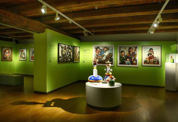 Ansicht eines Ausstellungsraums mit Gemälden an der Wand und einem Podest mit bunten Tonfiguren des Künstlers Michael Mathias Prechtl in der Mitte.