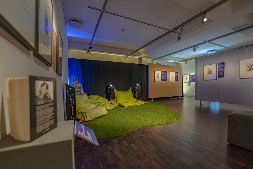 Ausstellungsraum mit Texttafeln, gerahmten Bildern und Sitzecke für Kinder mit hellgrünen Sitzsäcken auf einer Insel aus Kunstrasen.