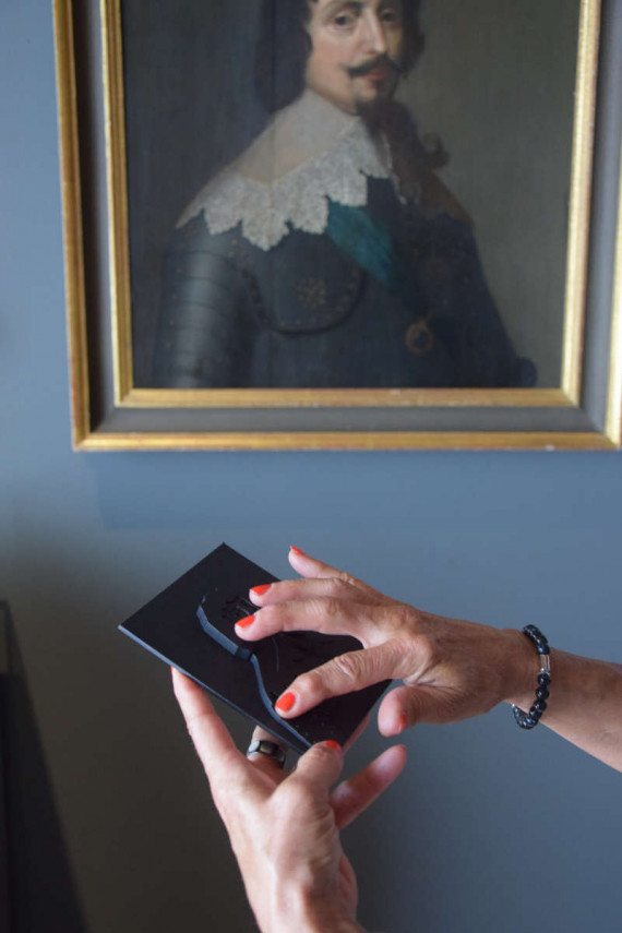 Zwei Hände Frauenhände ertasten ein kleines schwarzes Tastmodell. Im Hintergrund ist das Originalporträt zum Modell zu sehen, das Kurfürst Friedrich V. als Brustbild zeigt.