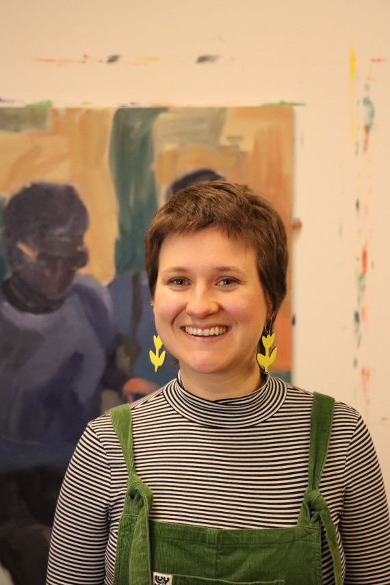 Frontales Brsutbild der Künstlerin Paula Schmidt im Atelier. Mit Kurzhaarschnitt, einem offenen Lachen, gelben Blumenohrhängern, einer dunkelgrünen Cord-Latzhose und einem weiß-schwarz gestreiften Shirt. 