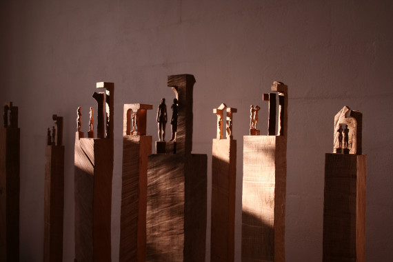 Querformatigers Bild mit Blick auf acht Holzstehelen mit kleinen figürlichen Holzskulpturen obendrauf.
