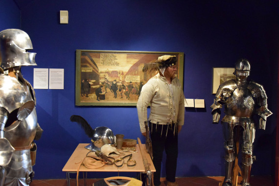 Mittelalterlich gekleidete Person neben einem Tisch zwischen zwei Ritterrüstungen stehend.