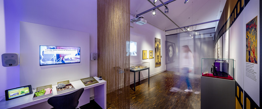 Blick in die Sonderausstellung mit einem Tisch und zwei Bildschirmen links, nach rechts öffnet sich der Blick in die Ausstellungen mit Tischvitrinen und hohen Textilwänden.