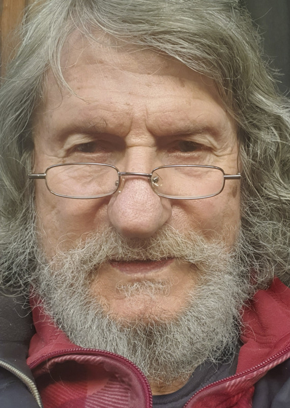 Frontales Porträtfoto vom Künstler mit kinnlangem grauem Haar und einem grauen Vollbart. Er trägt eine schmale Brille mit dünnem Gestell.