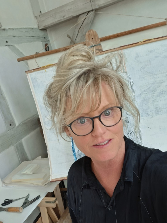 Brustbild einer blonden Frau mit Brille vor einer Tafel mit Landkarte