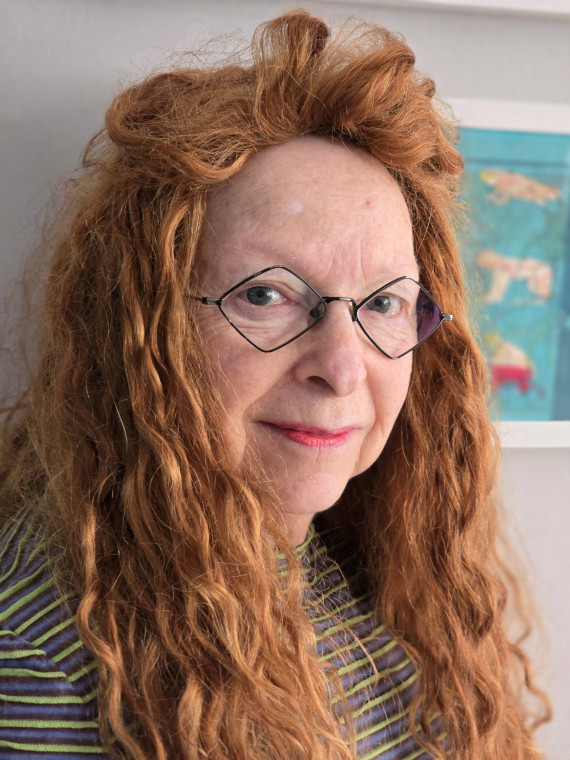 Brustbild im Dreiviertelprofil von einer Frau mit langem, kupferfarbenen und lockigem Haar und einer Brille mit rautenf&ouml;rmigen Gl&auml;sern. 