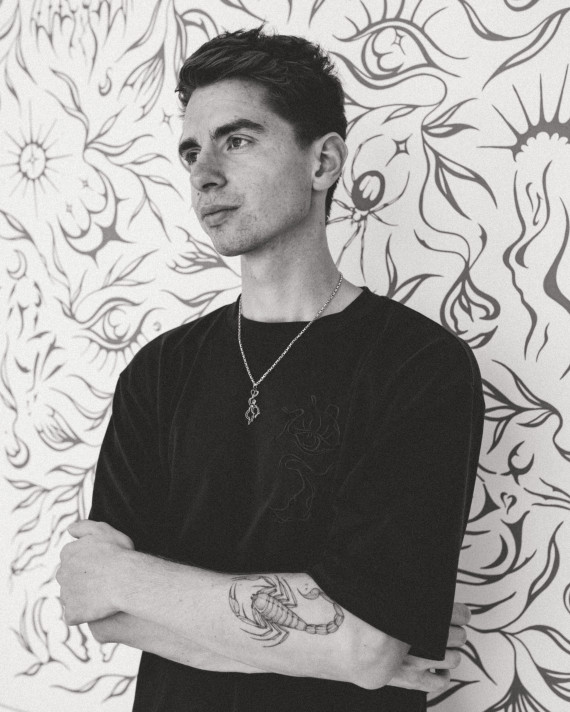 Schwarz-wei&szlig;es H&uuml;ftbild eines jungen Mannes in einem schwarzen T-Shirt. Der Blick geht nach links am Betrachter vorbei. Er steht vor einer gemusterten Wand.