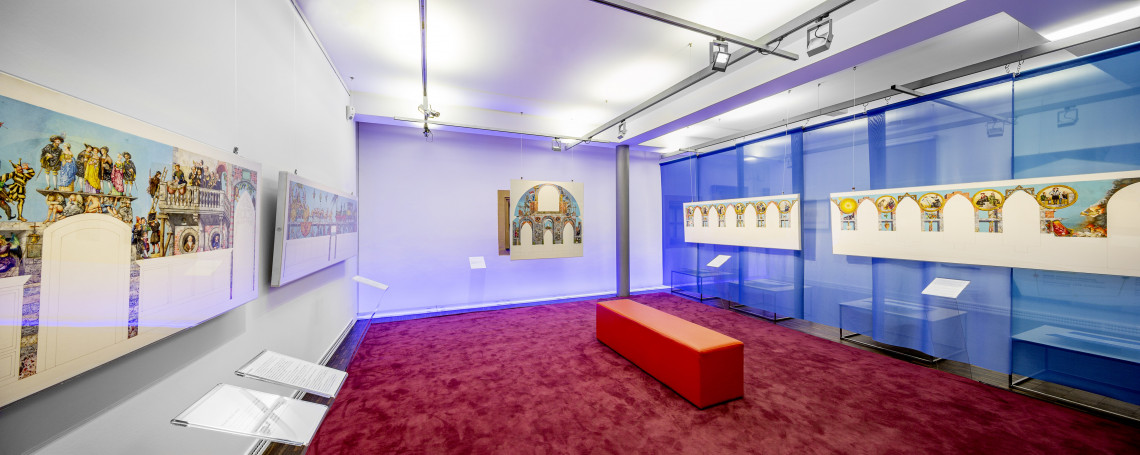 Ausstellungsraum mit mehreren Bildern an den W&auml;nden. Der Boden ist mit weinrotem Teppich ausgelegt. Mittig steht ein l&auml;nglicher roter Hocker.