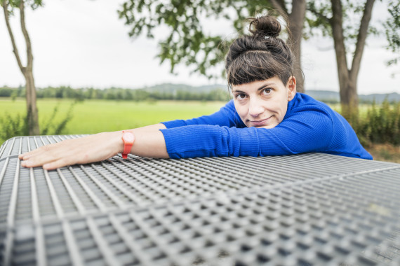 Eine Frau liegt mit Armen und Kopf auf einem Gittertisch und schaut direkt den Betarchter an. Sie tr&auml;gt die dunklen Haare in einem Dutt nach oben gebunden und hat einen Pony. Sie hat ein ultramarinblaues Oberteil mit langen &Auml;rmeln an und tr&auml;gt am Handgelenk eine kleine orangefarbene Uhr. Der Hintergrund &ouml;ffnet sich unscharf in eine weite Wiesenlandschaft mit vereinzelten B&auml;umen.